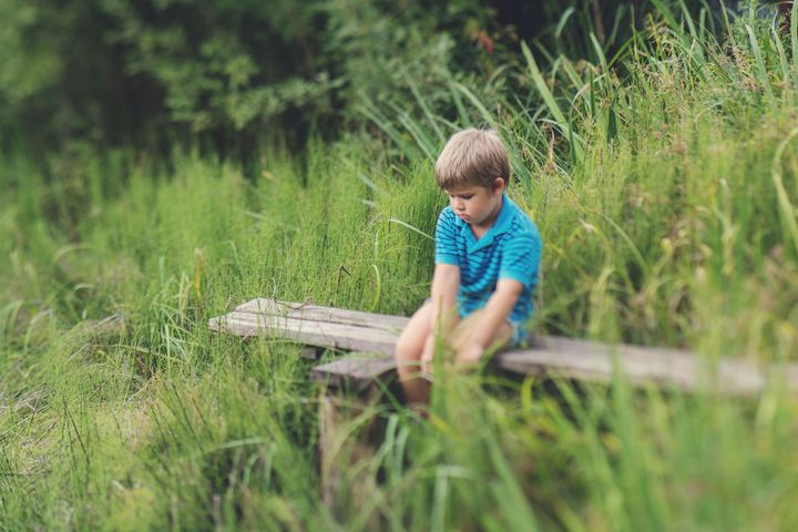 Jongen in een blauw shirt zit op een houten bankje in het hoge gras, buiten.