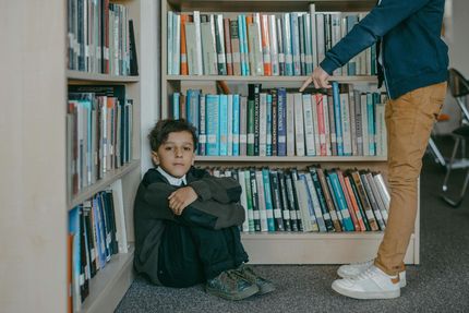 Een kind zit op de vloer van een bibliotheek tussen de boekenplanken, terwijl iemand anders naar een boek wijst.