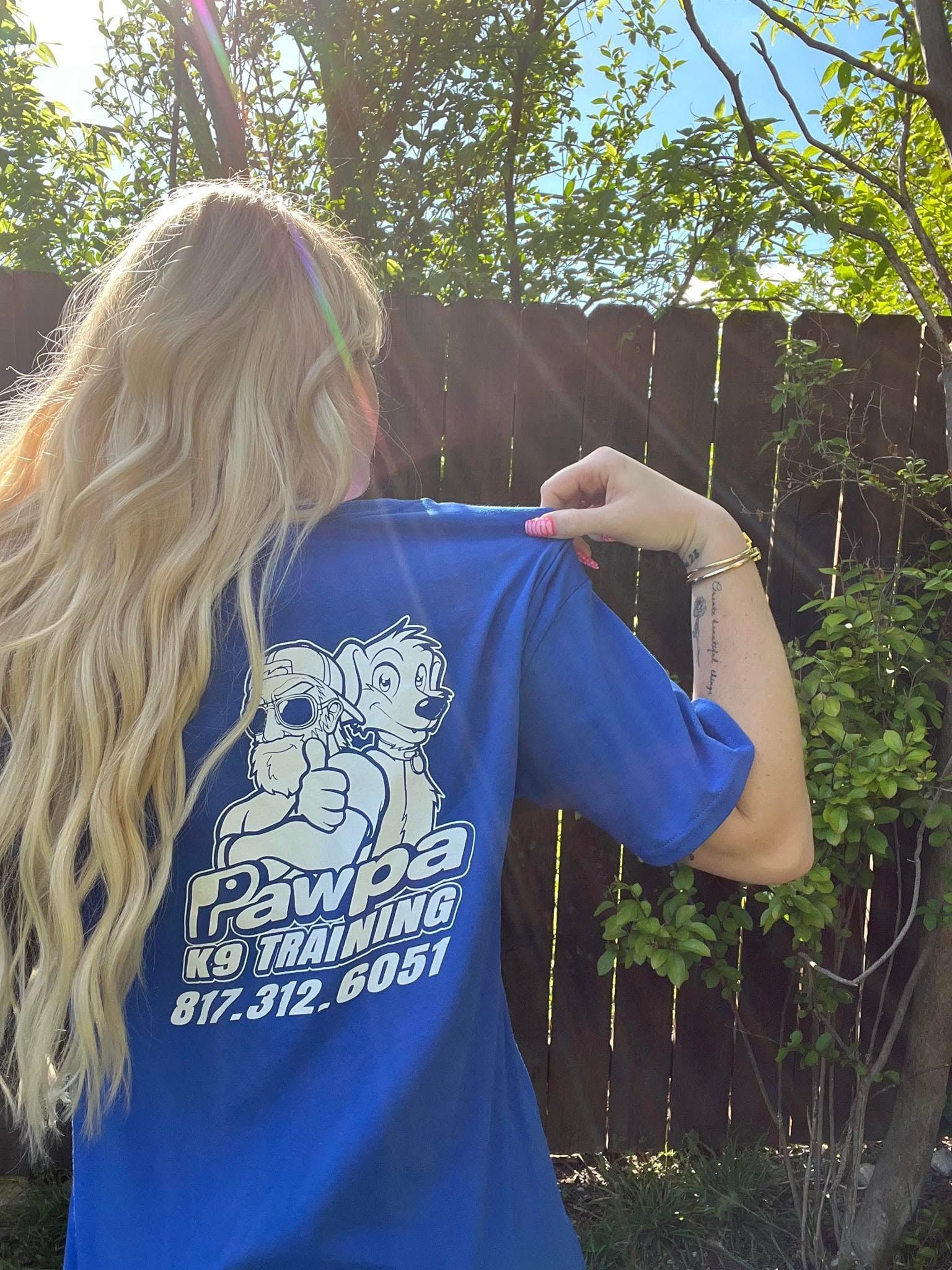 Person outdoors, showing the back of a blue T-shirt with a white graphic near a wooden fence.