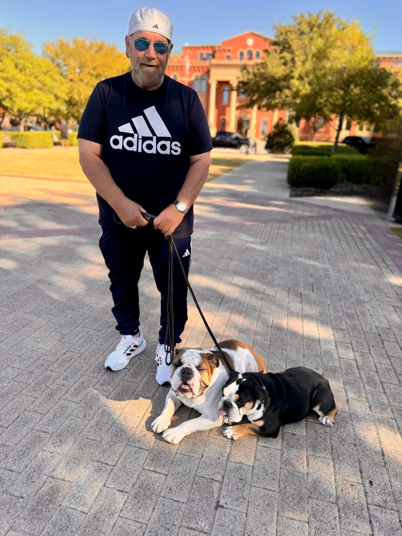 Man standing with two dogs on a sunny walkway in front of a building, autumn trees in the background