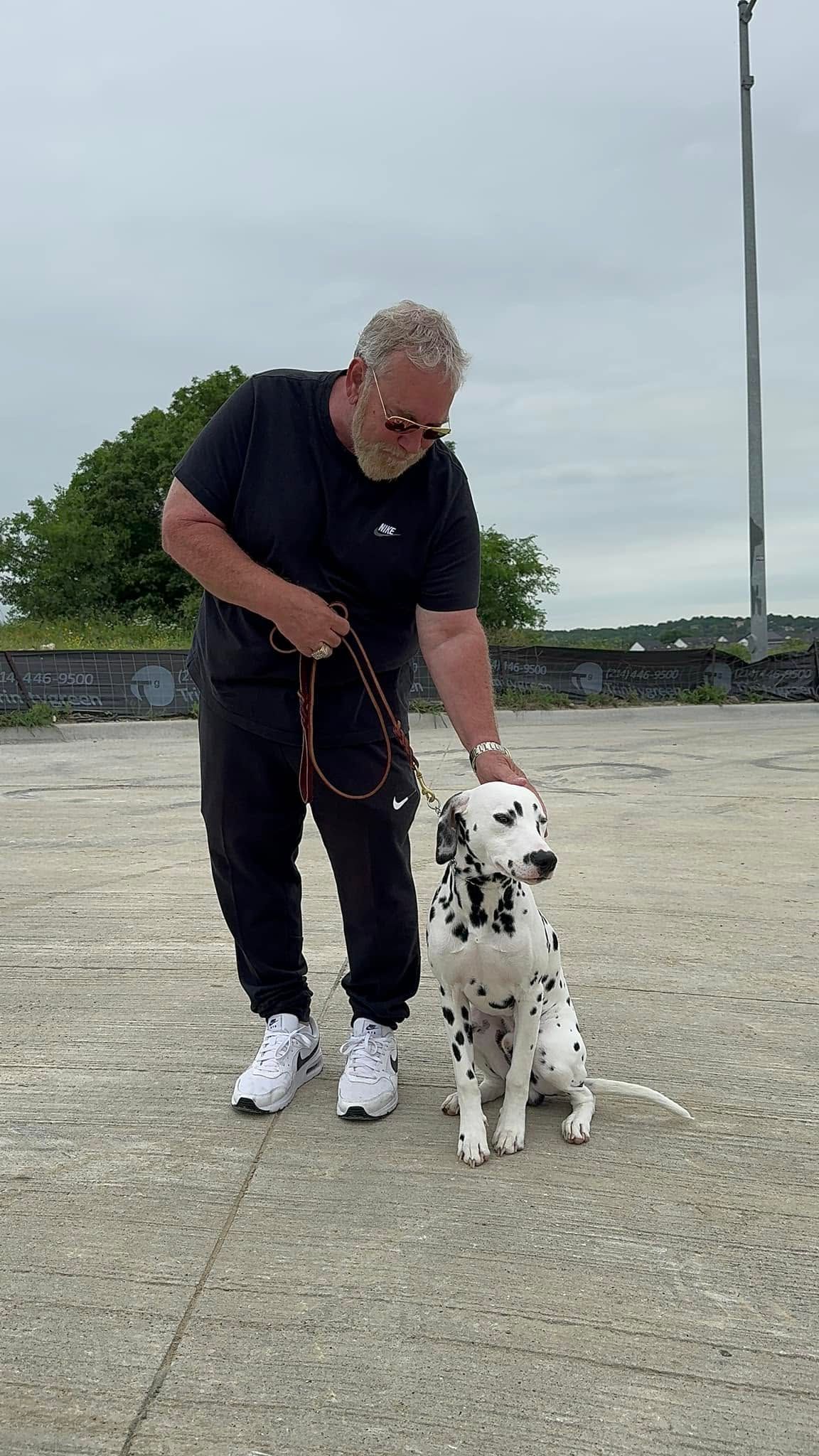 Man in dark clothes bending beside a Dalmatian on a leash outdoors on a cloudy day