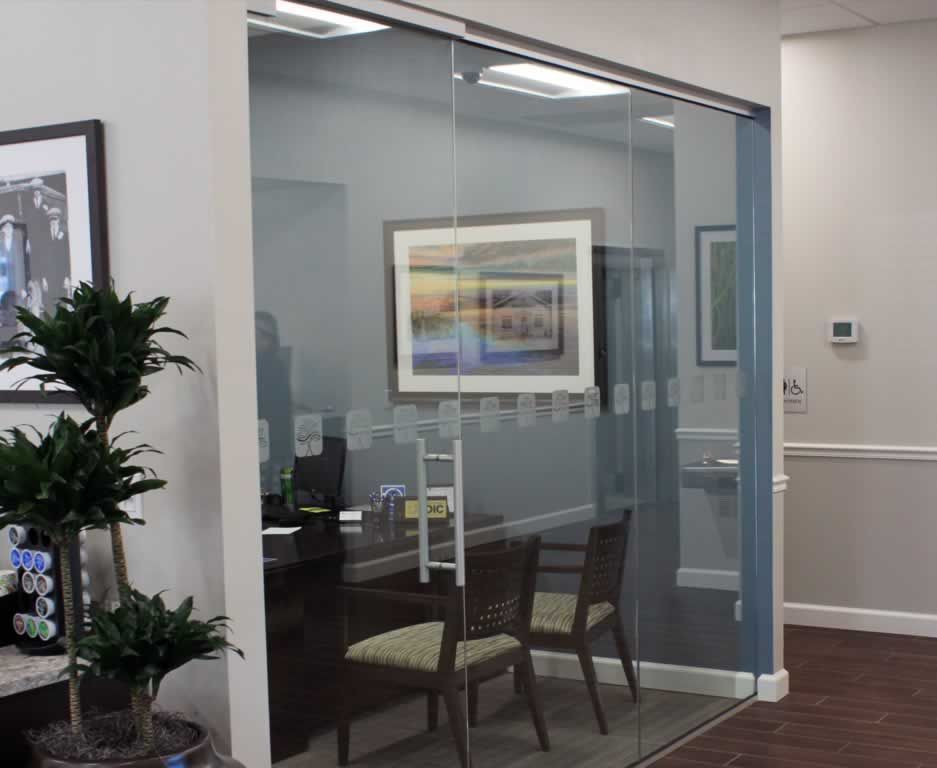 Glass-walled office meeting room with framed artwork, chairs, and a potted plant nearby