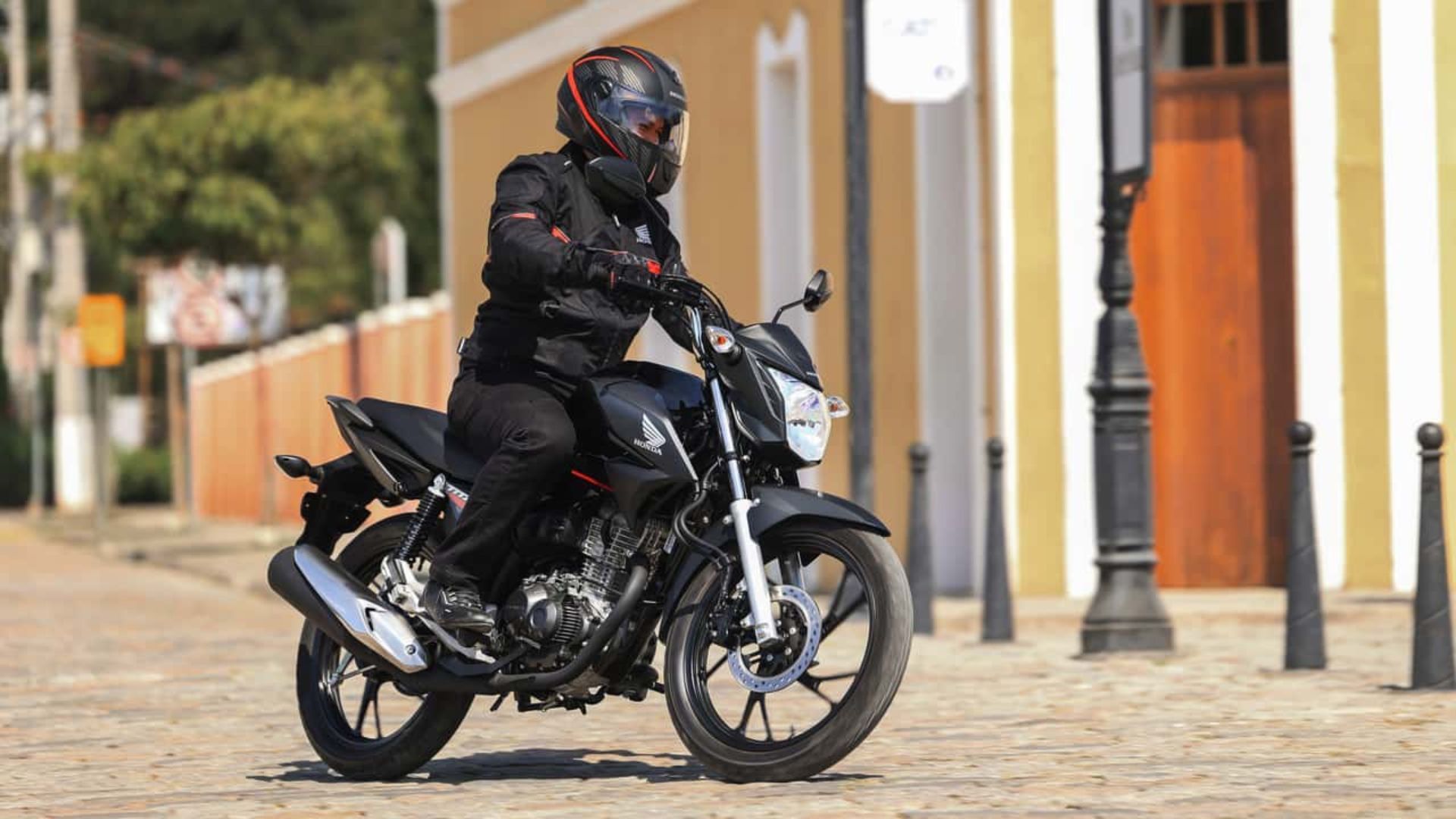 Motorcyclist in black gear riding a black motorcycle on a cobblestone street beside orange buildings