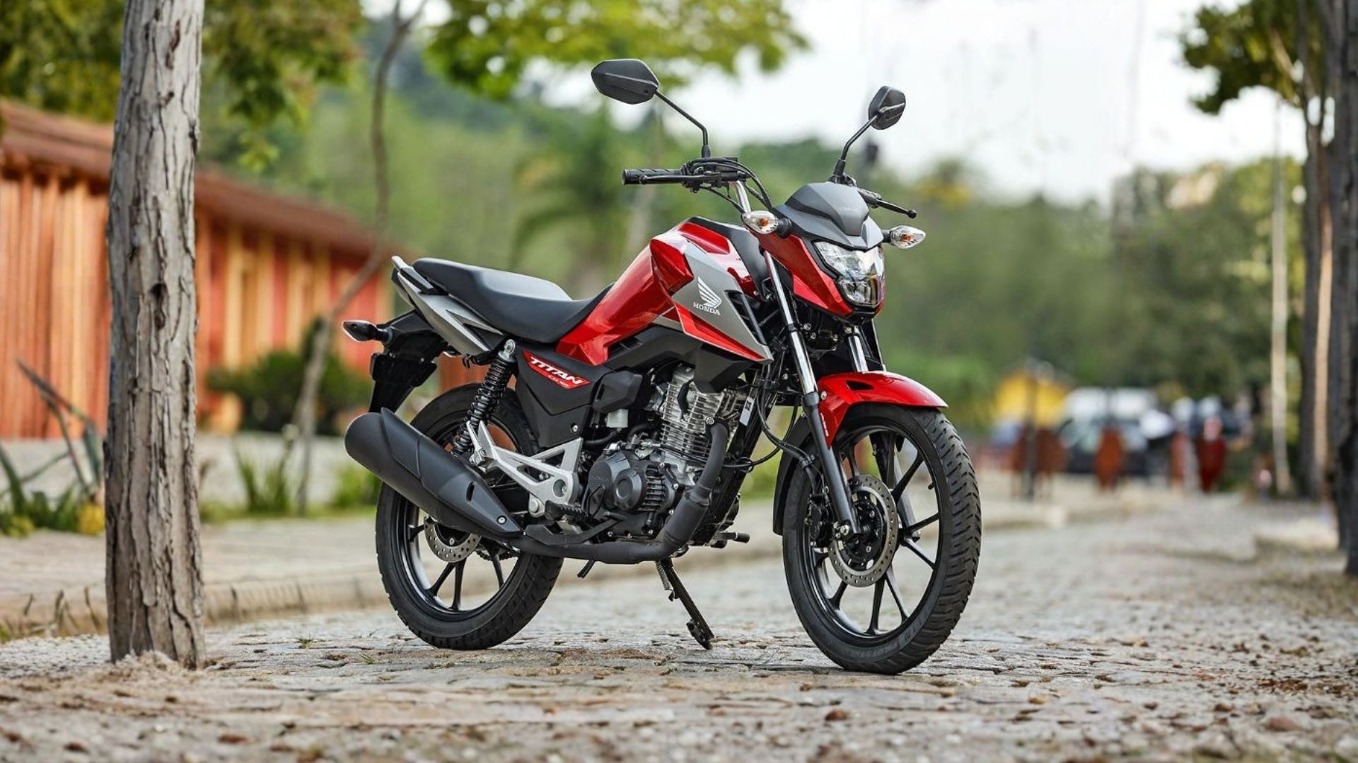 Red motorcycle parked on a cobblestone street near a wooden fence, with trees and people in the background
