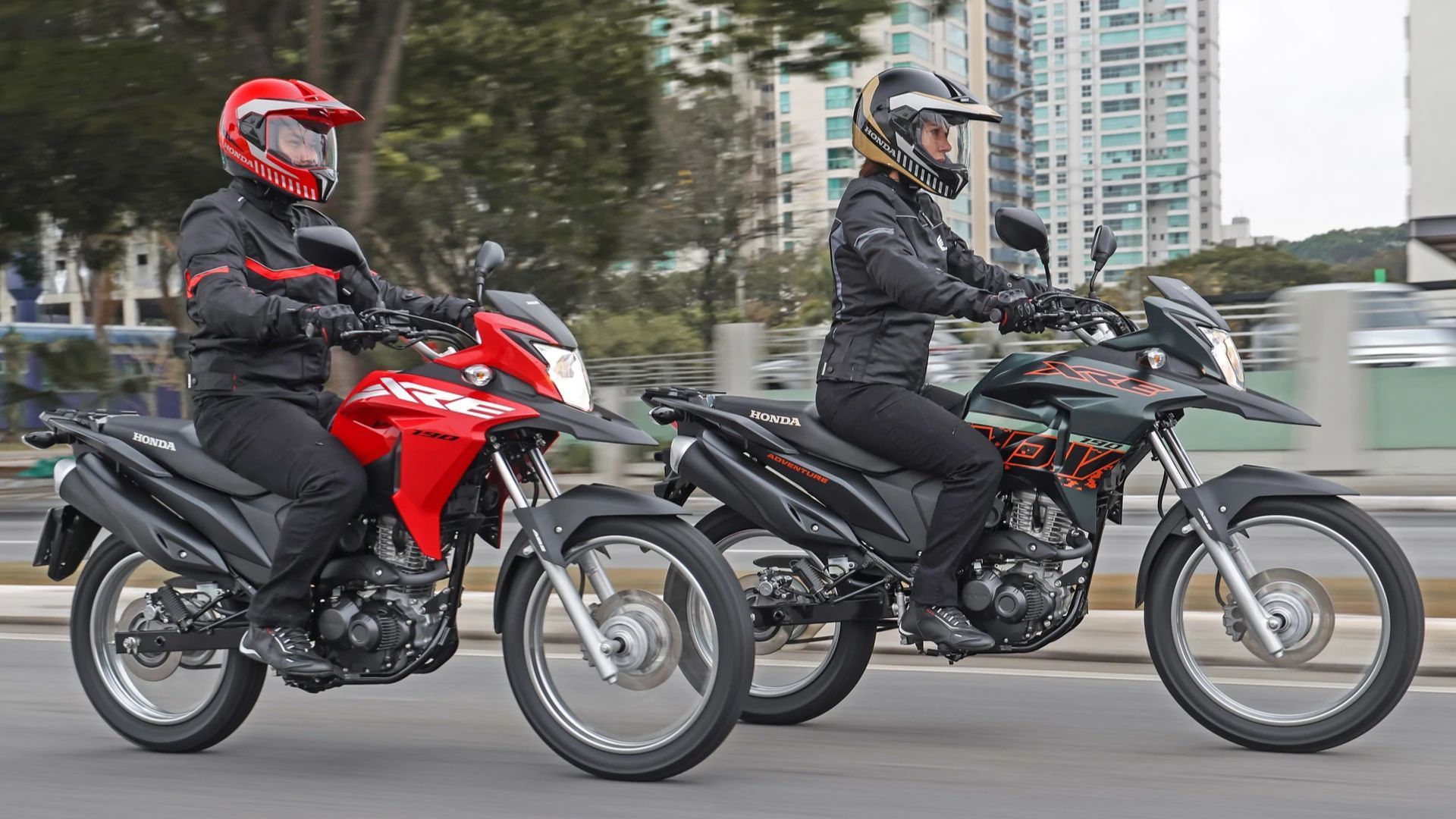 Two motorcyclists riding side by side on a city street, one on a red bike and one on a black bike.