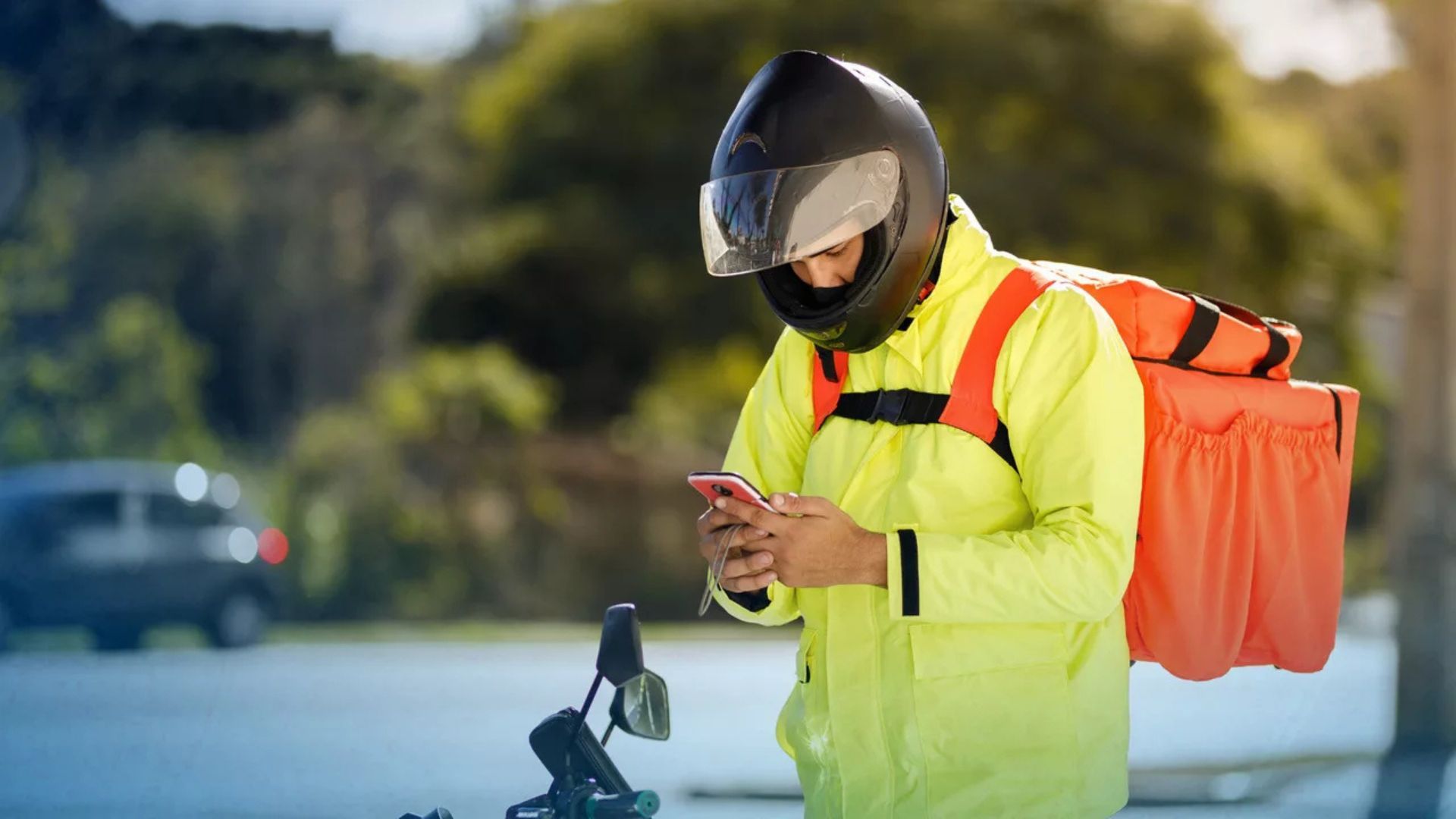 Delivery worker in neon jacket and helmet checking a phone beside an orange backpack.