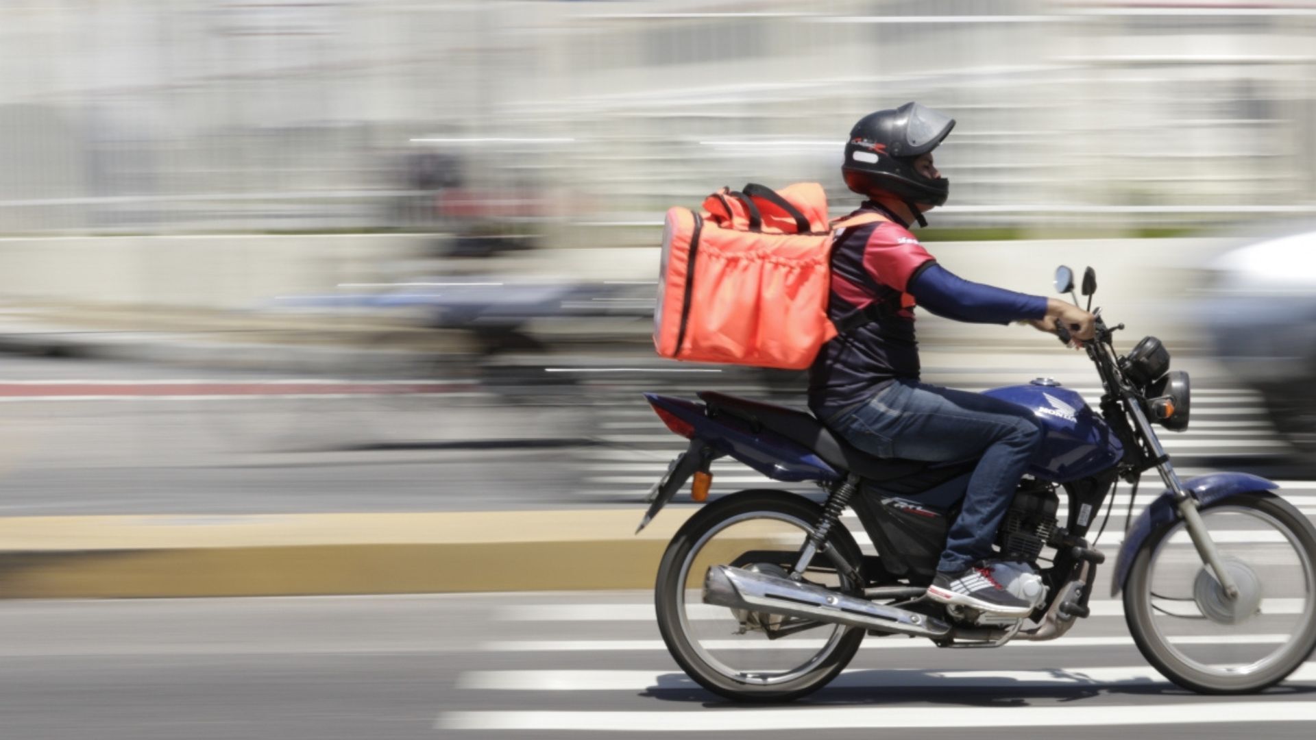 Delivery rider on a motorcycle with a red backpack, speeding along a city street.