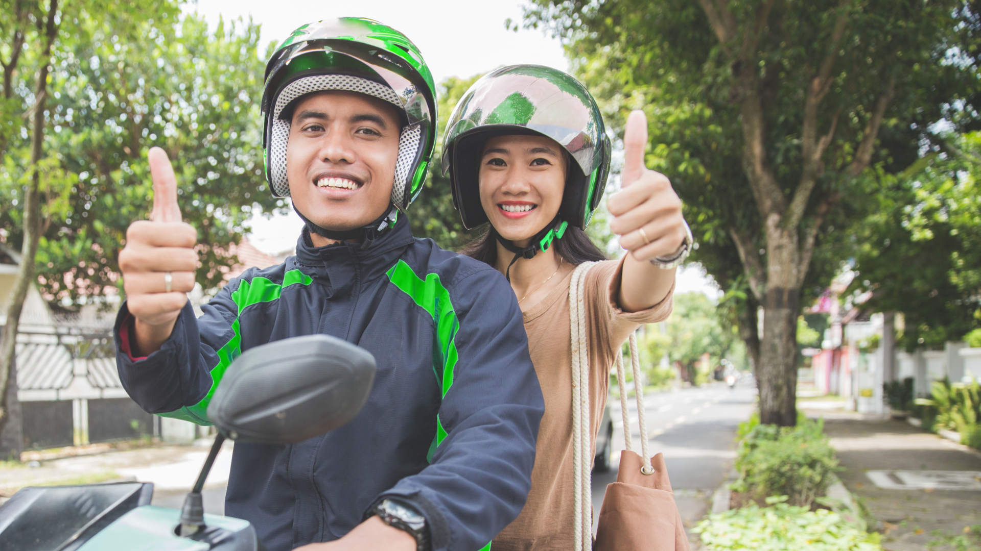 Two people on a scooter smiling and giving thumbs up on a tree-lined street