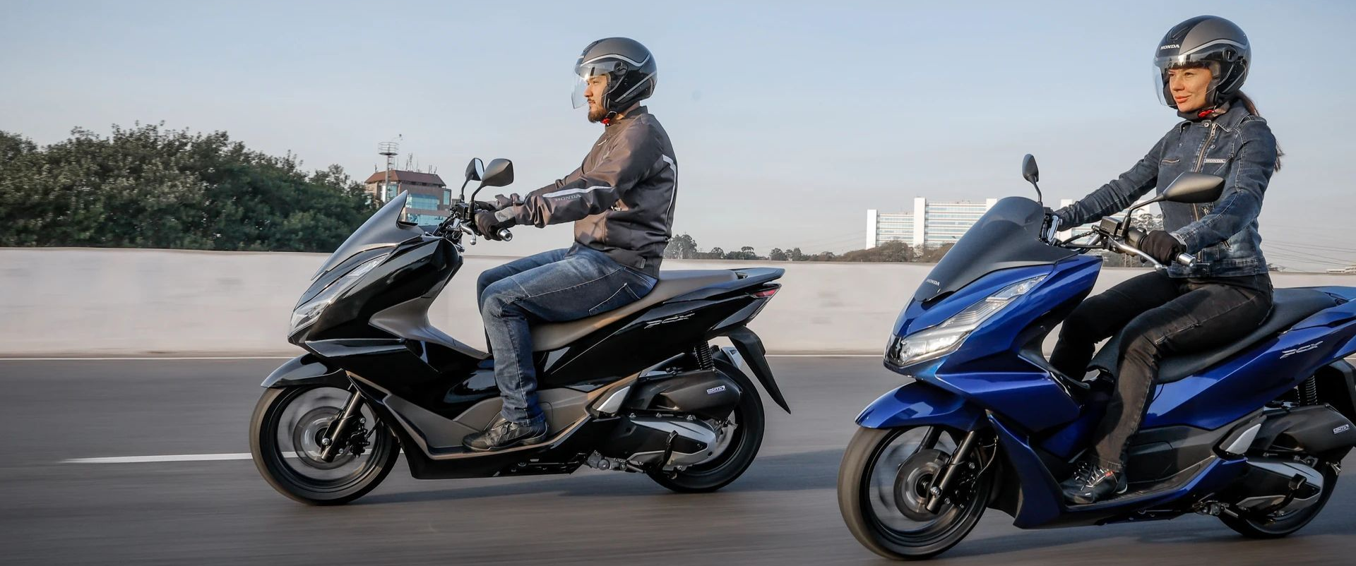 Two helmeted riders on scooters, one silver and one blue, ride side by side on a road.