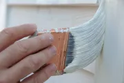 Hand holding a paint brush, applying white paint to a wooden surface.