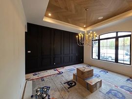 Room with dark panel wall, parquet ceiling, large window, and chandelier. Construction materials on floor.