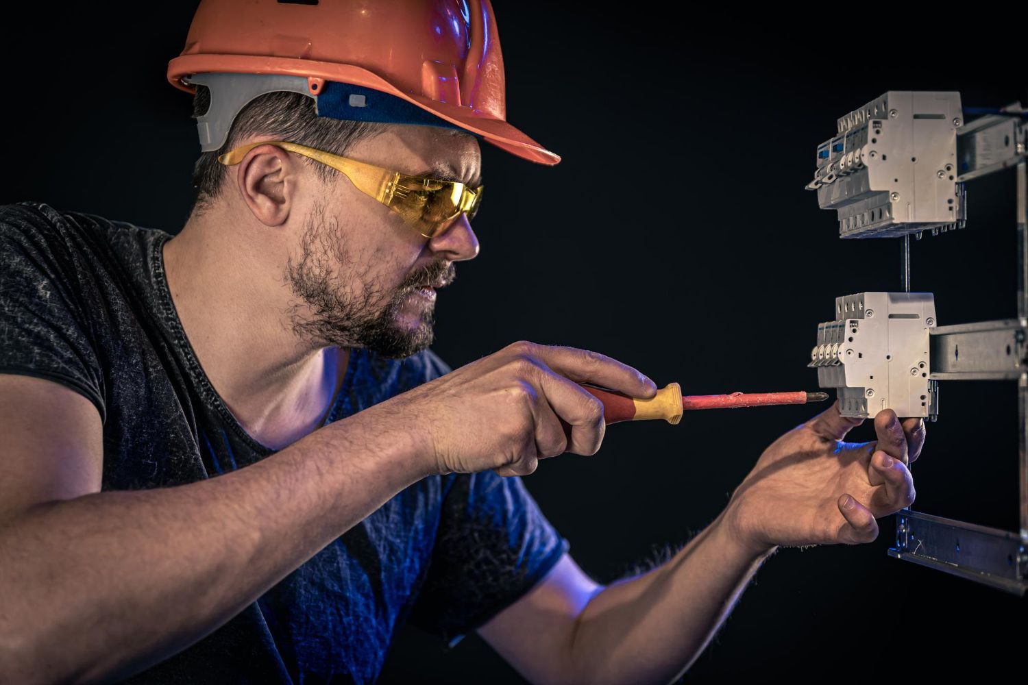 Electrician in an orange hard hat and safety glasses using a screwdriver on electrical equipment.