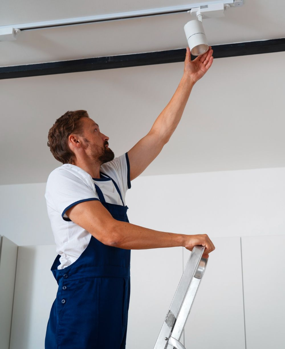 Man on a ladder installing a track light fixture on a ceiling.