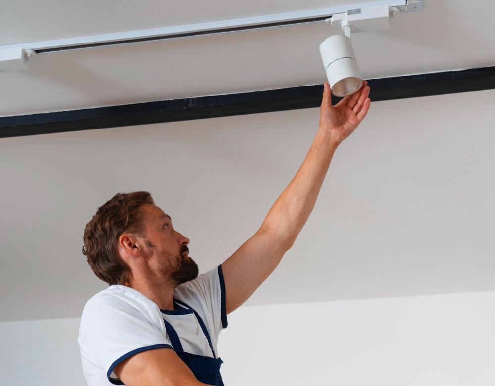 Man installing a spotlight on a track lighting system. White ceiling, black beam, he’s wearing a blue apron.