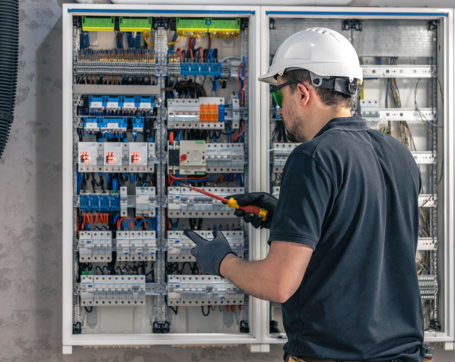 Electrician in a hard hat working on an electrical panel with a screwdriver.