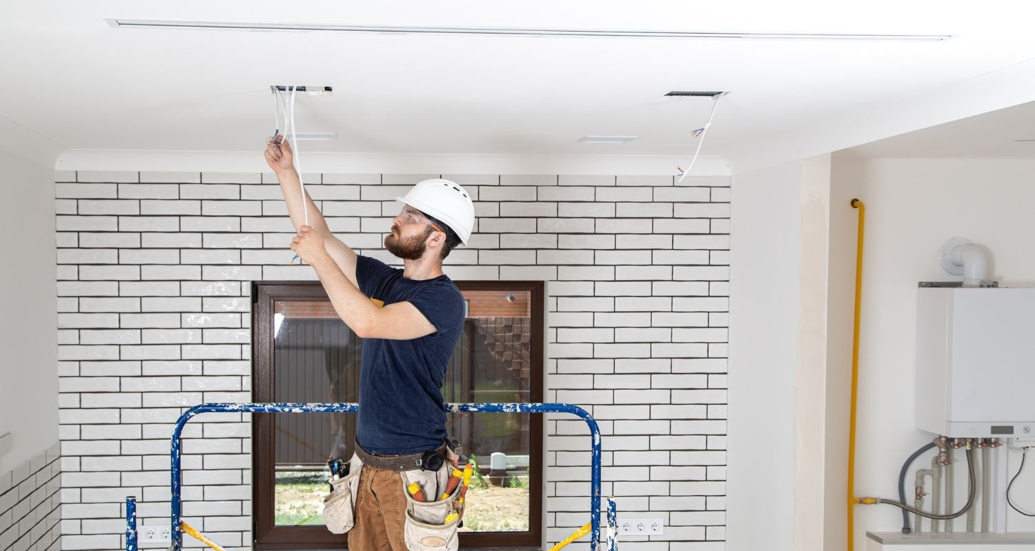 Electrician on a ladder installing wiring in a room with white brick wall and a gas boiler.