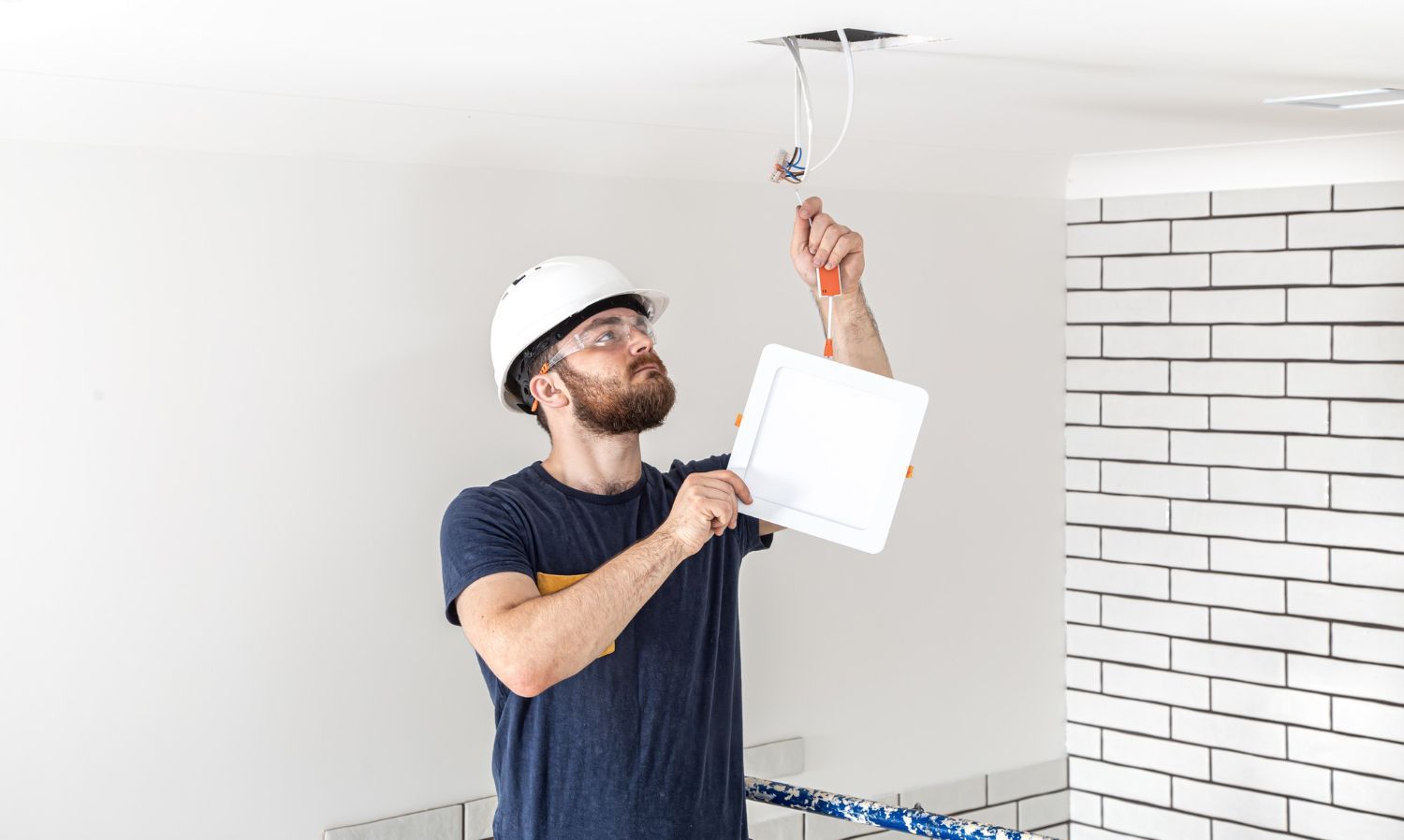Electrician in a hard hat inspecting wiring in a ceiling, holding a plan in a white room.
