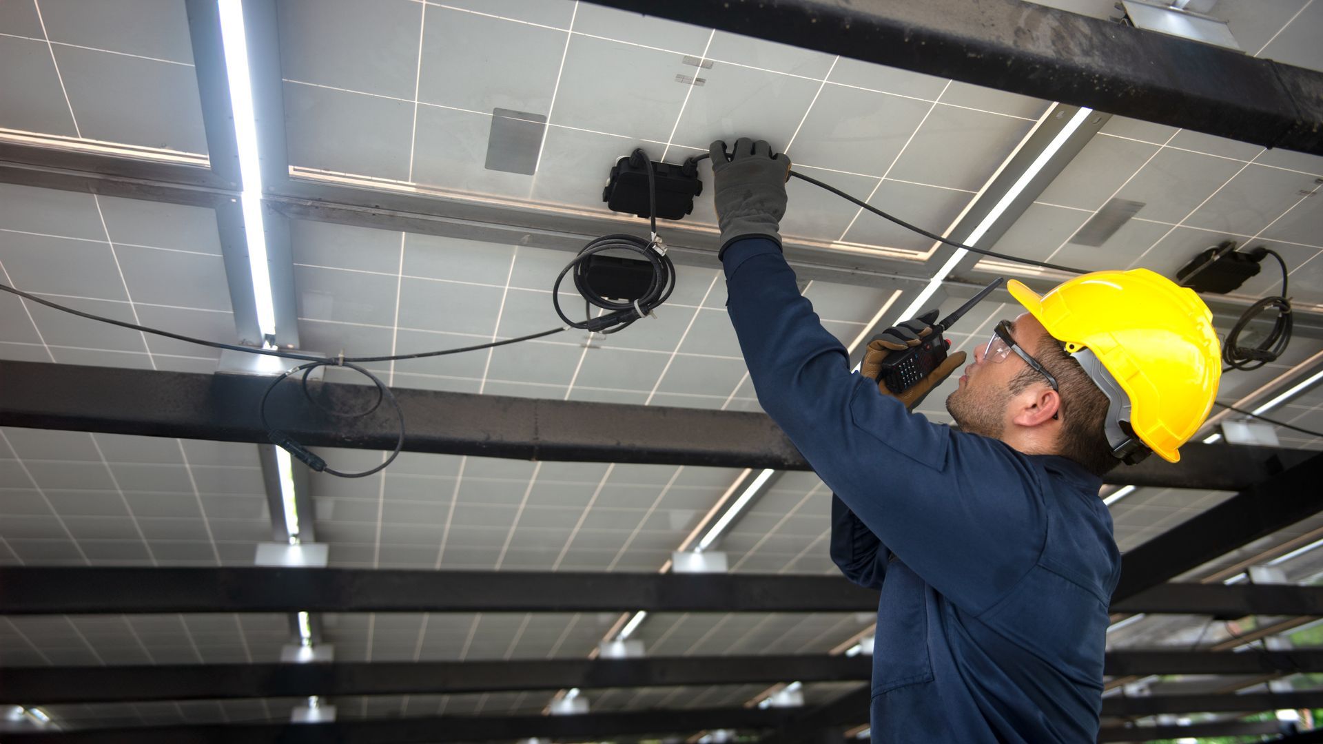 Solar panel technician in yellow hard hat connecting wires under solar panels.