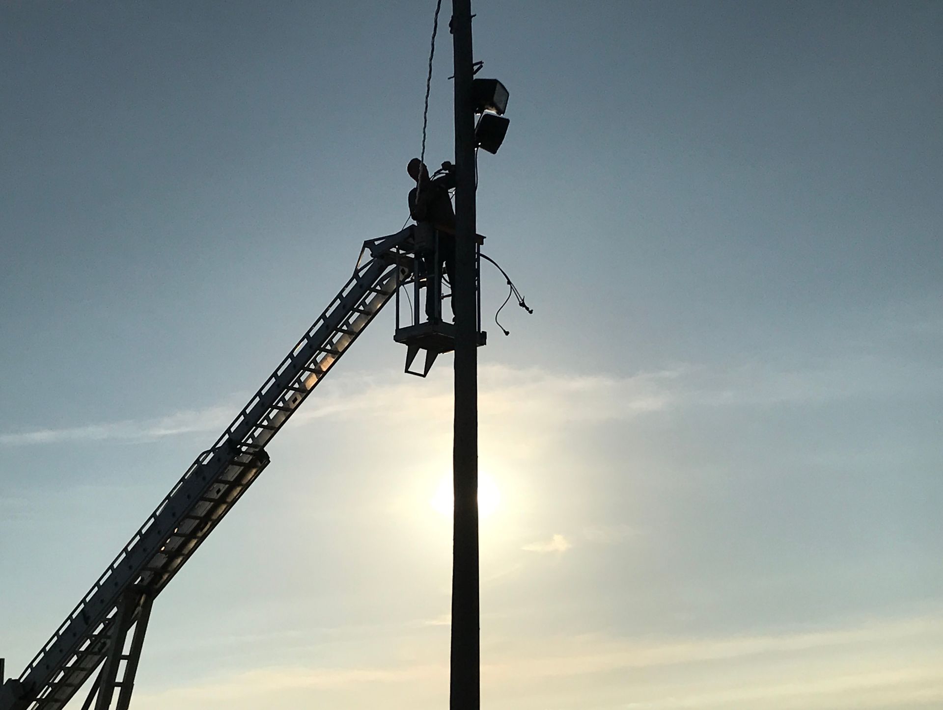 Silhouette of workers in a bucket lift on a tall pole, silhouetted against a bright sun in a blue sky.