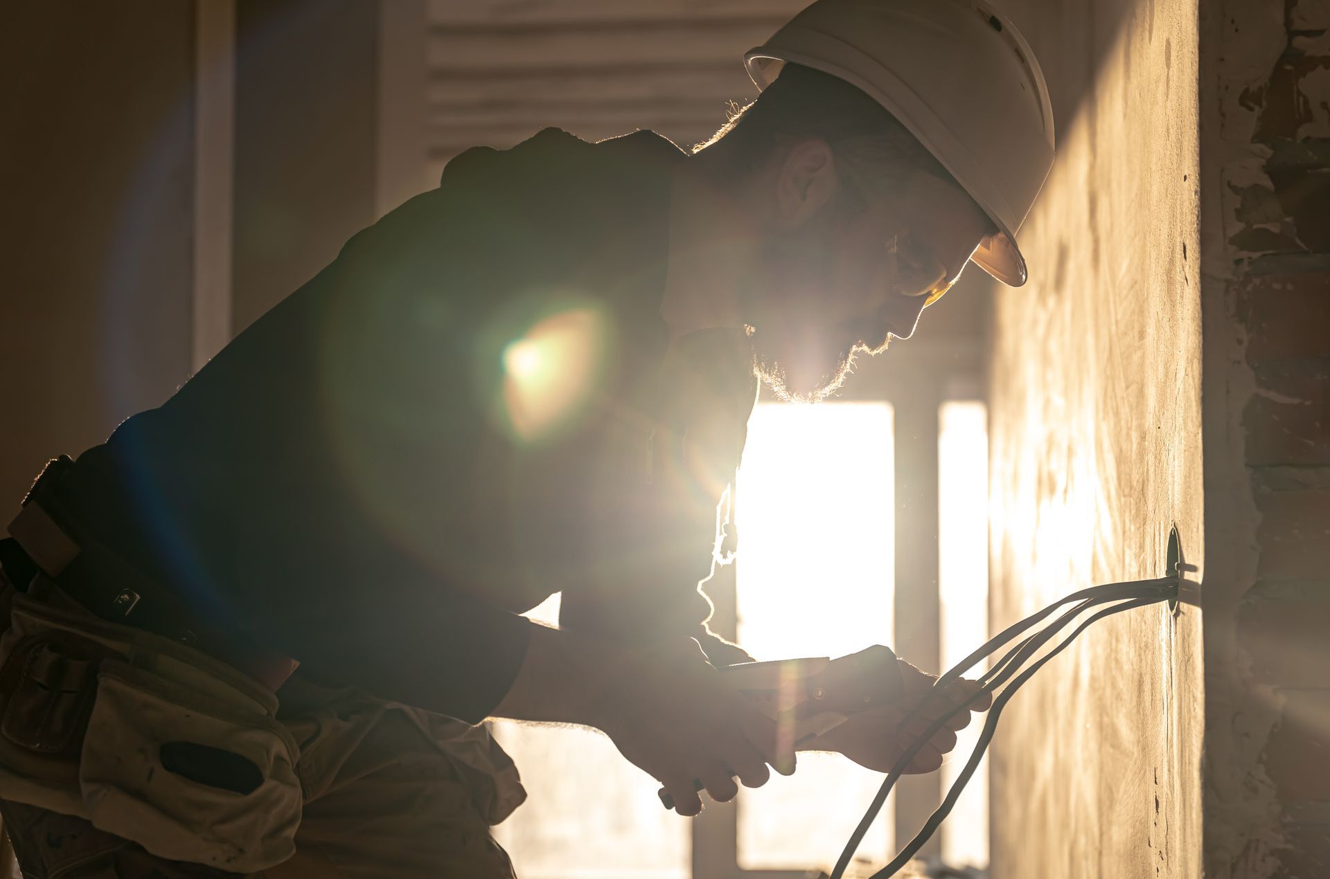 Electrician wiring an outlet in a sunlit room, wearing a hard hat and tool belt.
