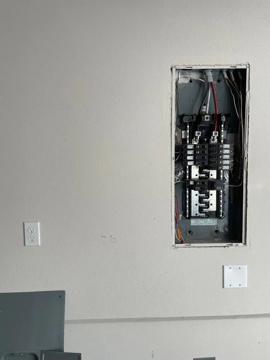 Electrical panel recessed in a light gray wall with wiring exposed. Two white electrical outlets are visible.