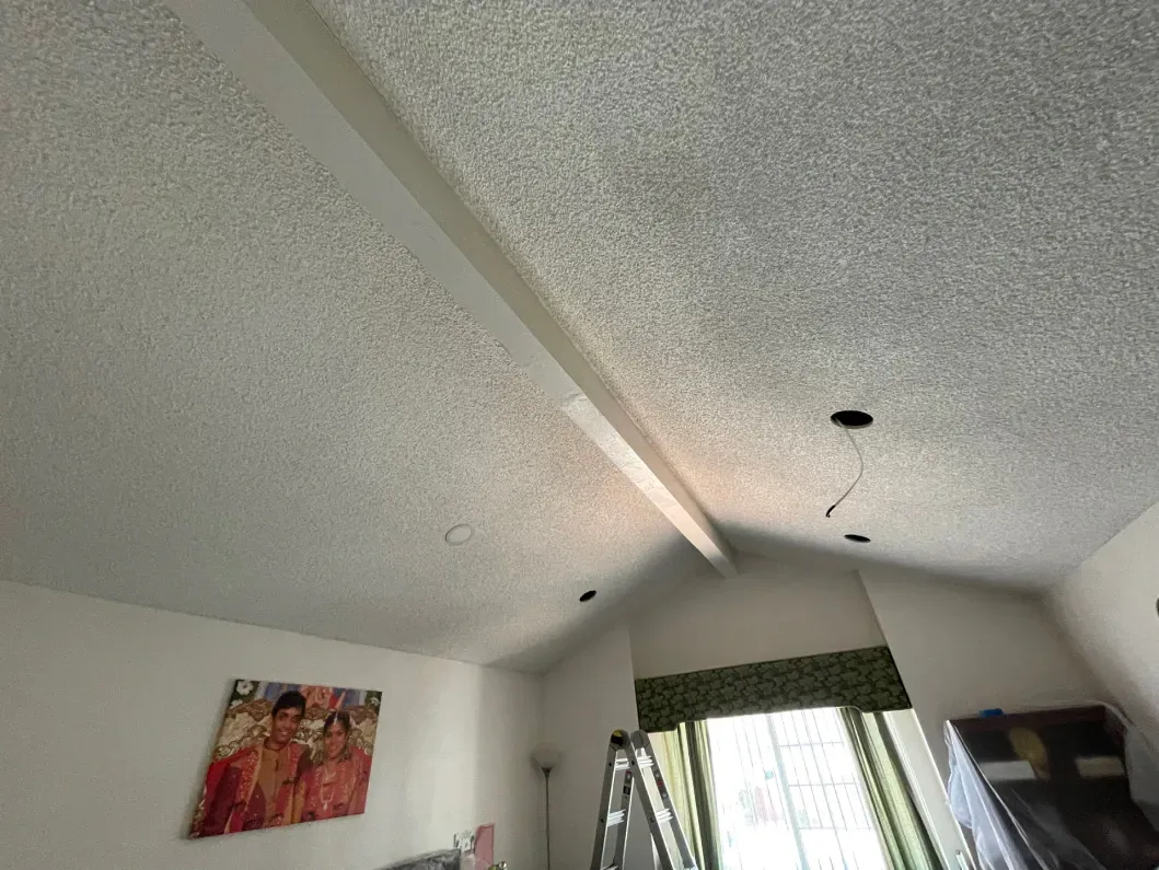Textured ceiling with a central beam, white walls, and a window with green curtains.