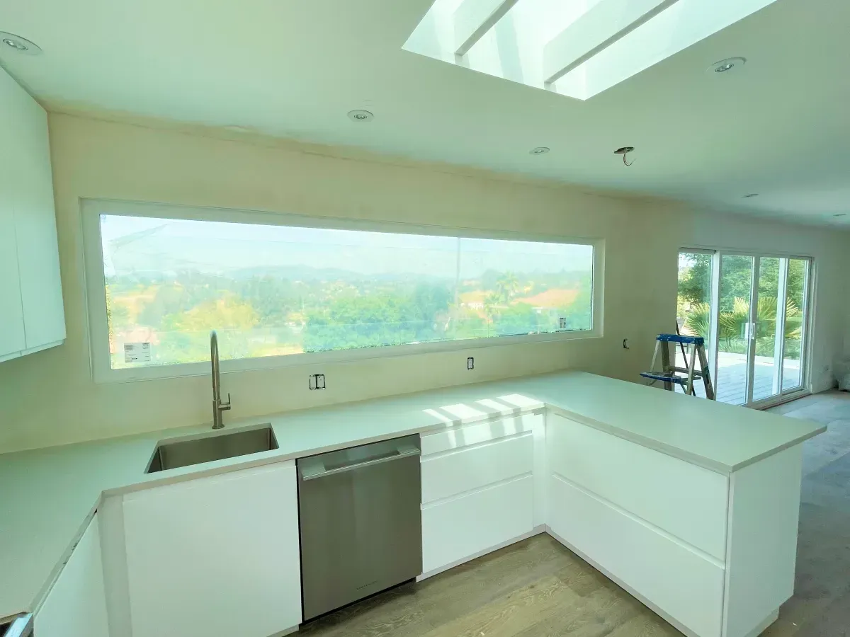 Modern white kitchen with large window overlooking greenery and a skylight. Stainless steel sink and dishwasher.