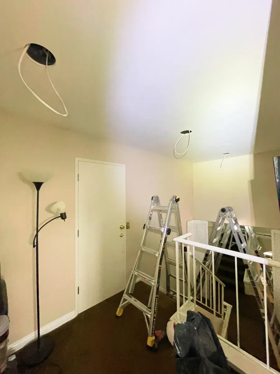Ceiling with exposed wiring, door, ladder, floor lamp, and a cluttered room.