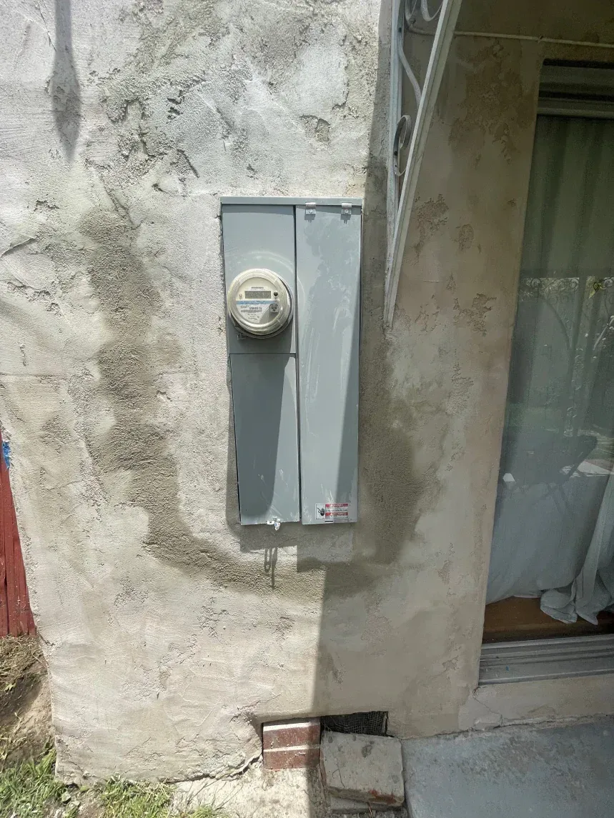 Electric meter and panel mounted on a weathered stucco wall next to a window.