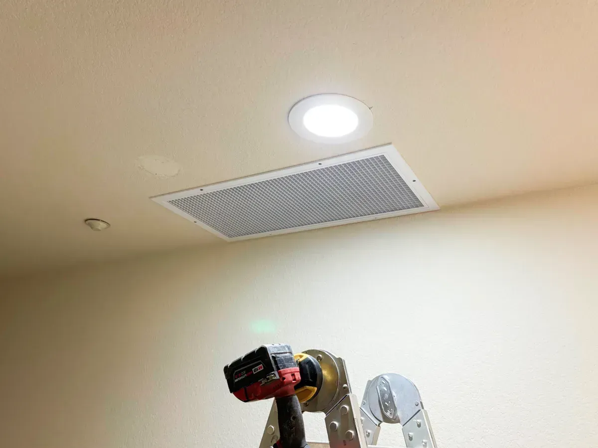 Ceiling with recessed lights and large air vent. A person is working on a ladder with a power drill.