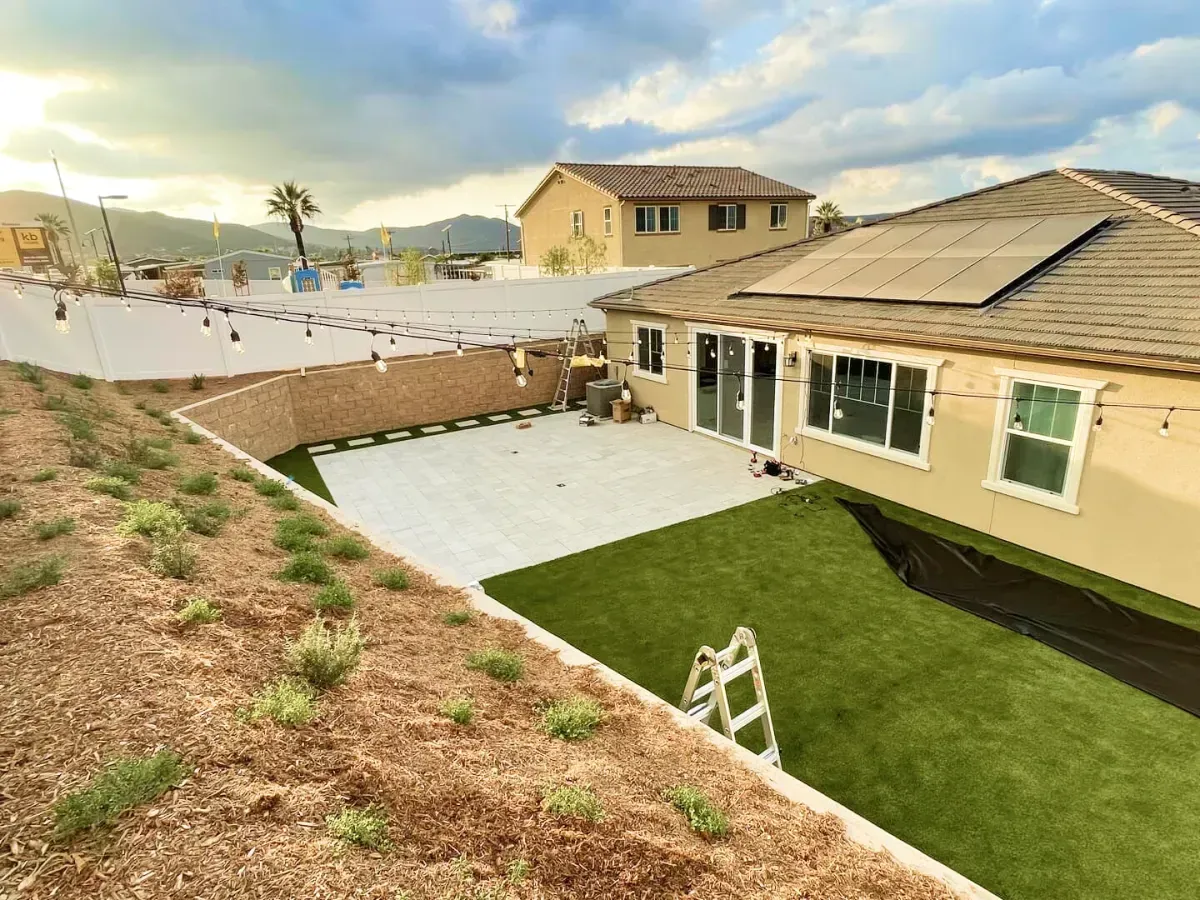 Backyard with beige house, patio, and artificial grass. White fence, string lights, and a ladder are visible.