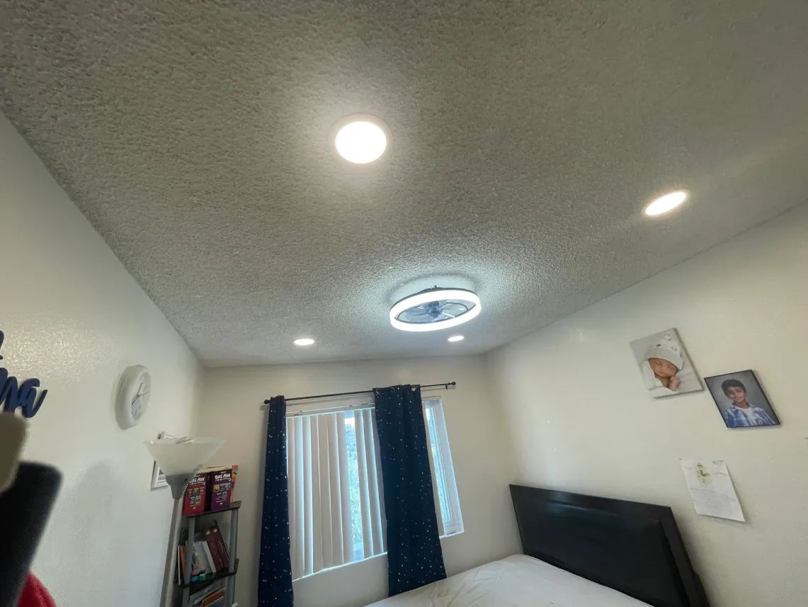 A bedroom ceiling with a light fixture and recessed lights. A window and bed are below.