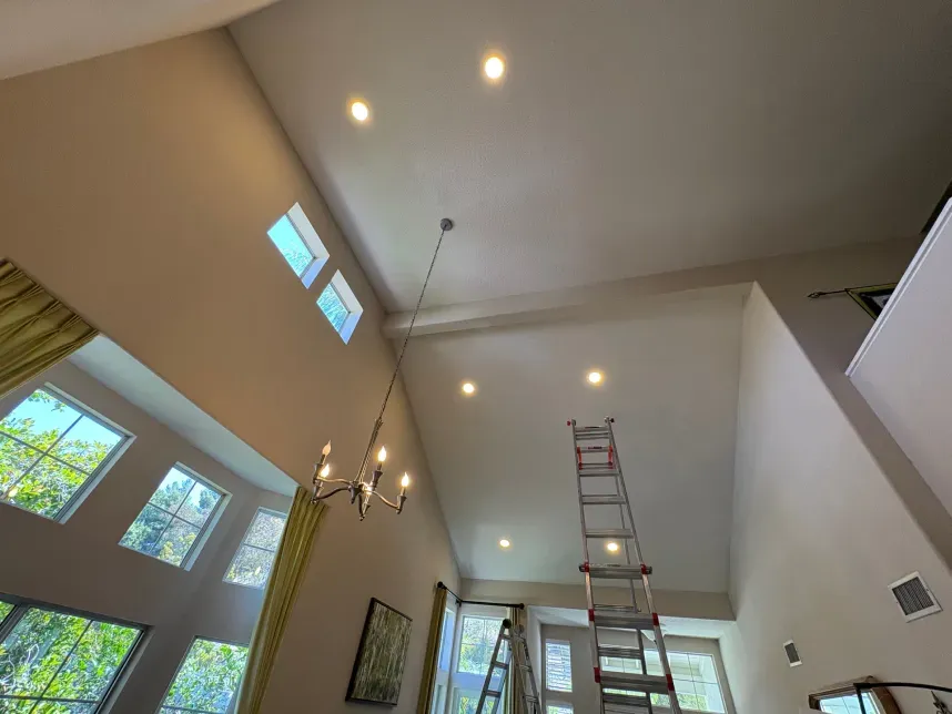 High-ceilinged living room with recessed lights. Windows on the left, chandelier hanging, ladder set up in the center.