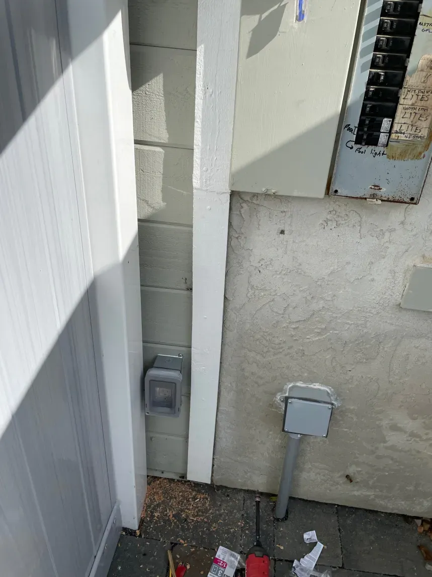 A white electrical conduit runs vertically along a cinder block wall, with a breaker box and electrical outlets.