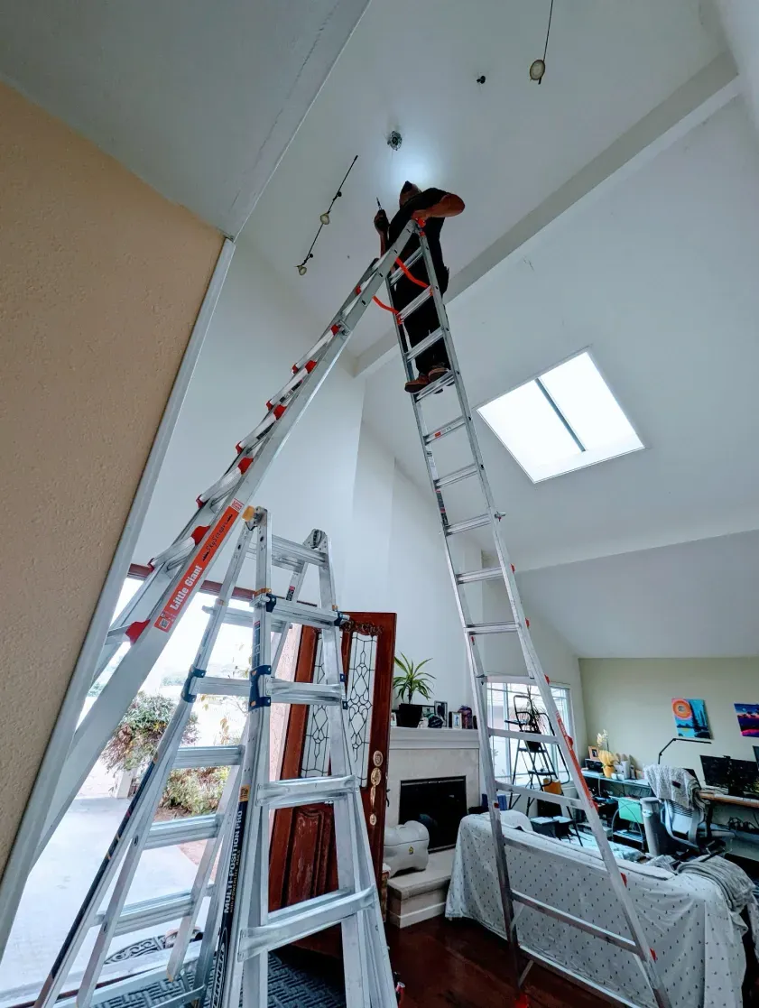 Person on ladders working on ceiling lights in a room with a fireplace and skylight.