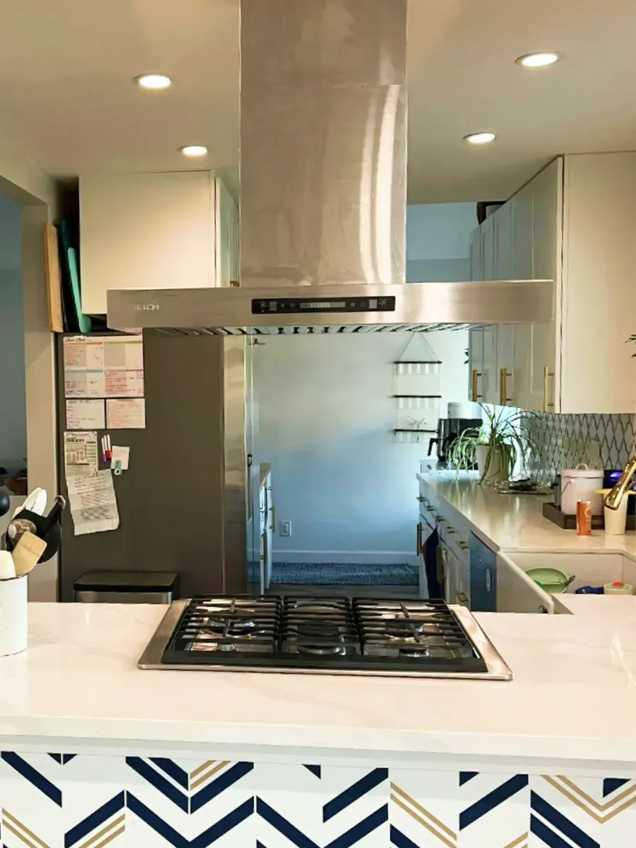 Modern kitchen with stainless steel range hood above a gas cooktop, white countertops, and chevron tile backsplash.