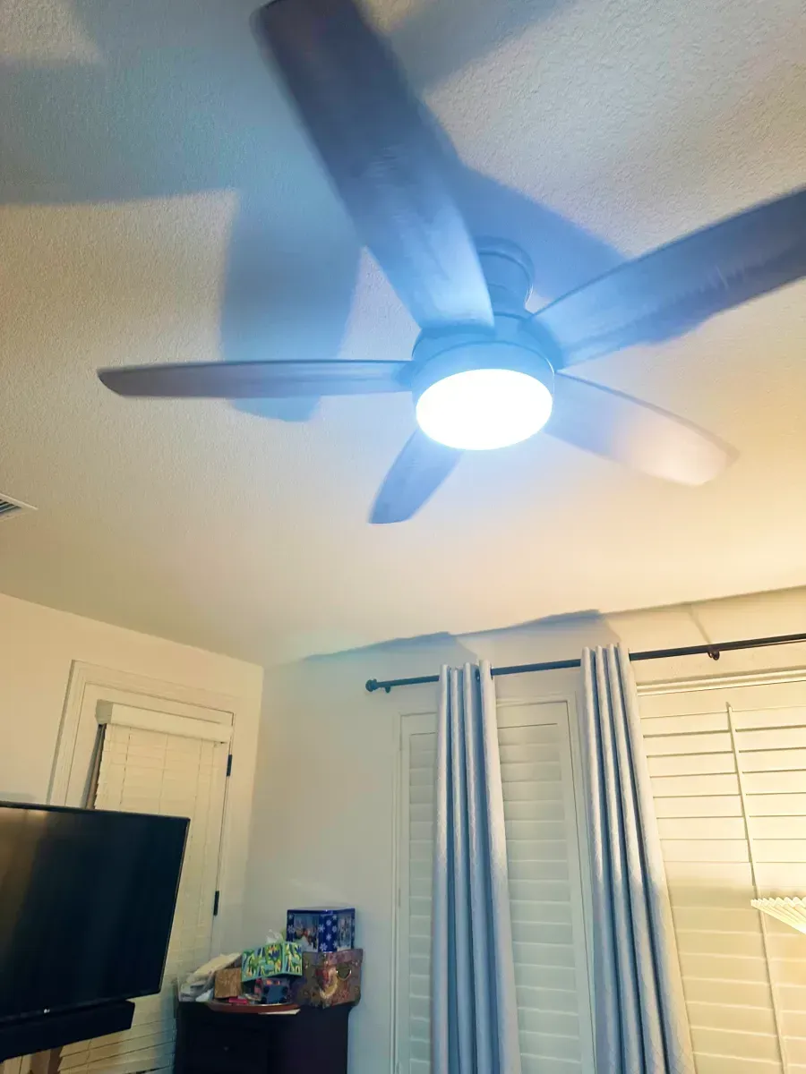Ceiling fan with light fixture illuminates room; window with curtains and blinds, TV visible.