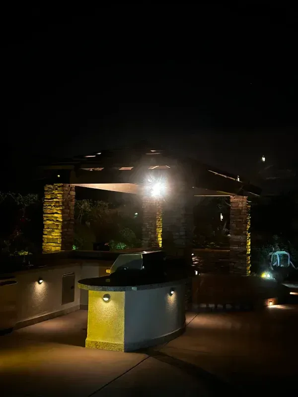 Outdoor kitchen at night, lit by warm lights. Structure with stone facade and grill.