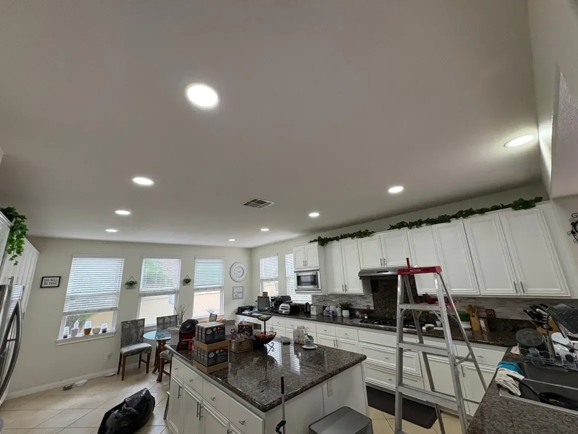 Bright kitchen with white cabinets, island, and ceiling lights; windows and appliances visible.