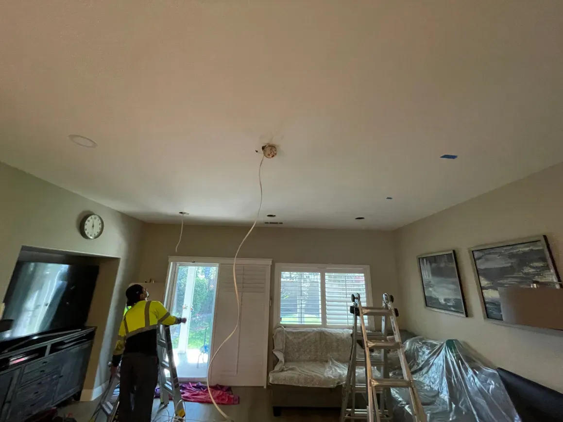 A person on a ladder installs wiring in a living room ceiling. Wires hang down near the center.