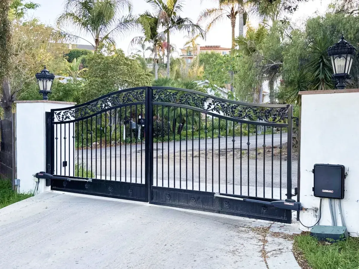 Black wrought-iron driveway gate, white pillars, street lamps. Gate is closed. Palm trees in the background.