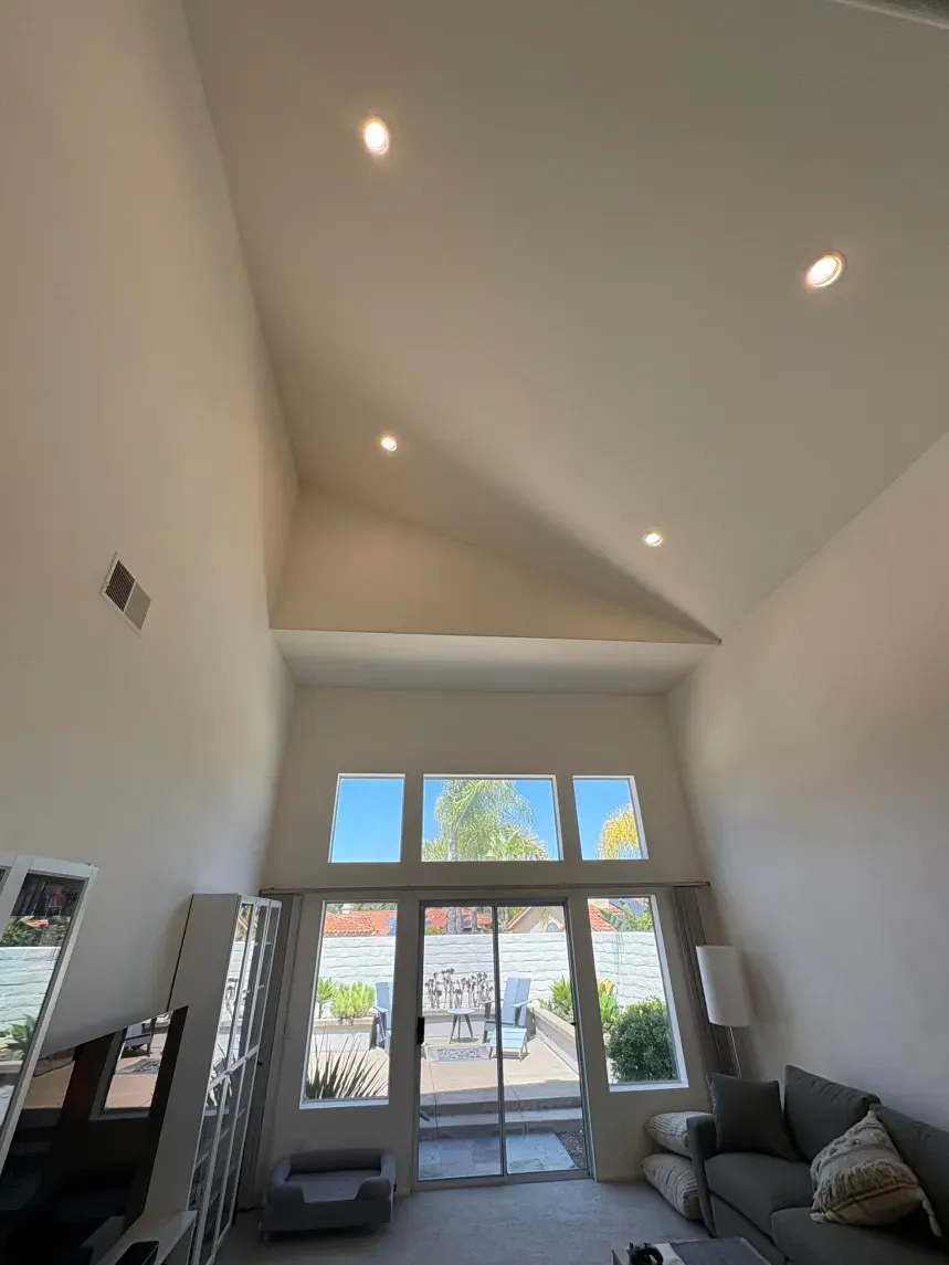 High-ceilinged living room with a sliding glass door and windows. Recessed lighting. Gray couch, white walls.