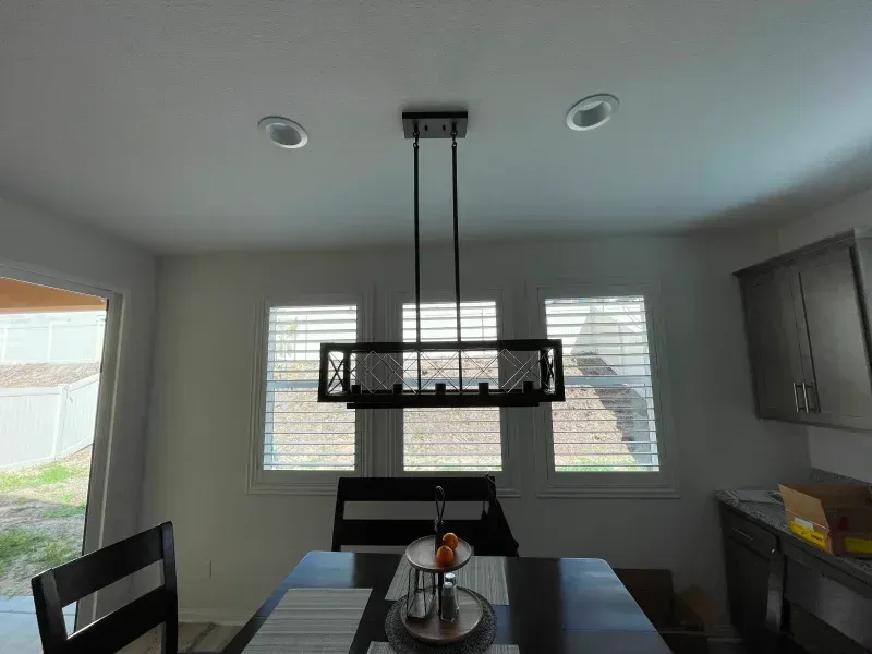 Dining room with a black chandelier over a dark wood table set near windows with blinds.
