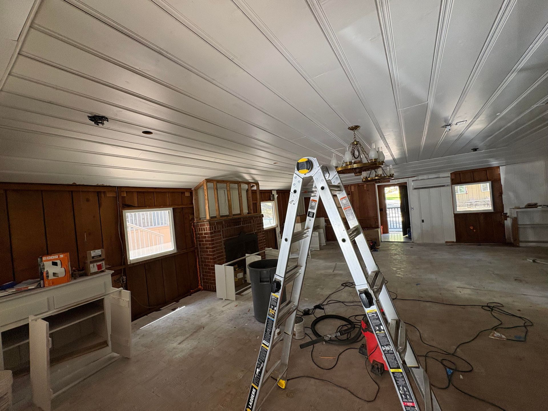 Person on ladders working on ceiling lights in a room with a fireplace and skylight.