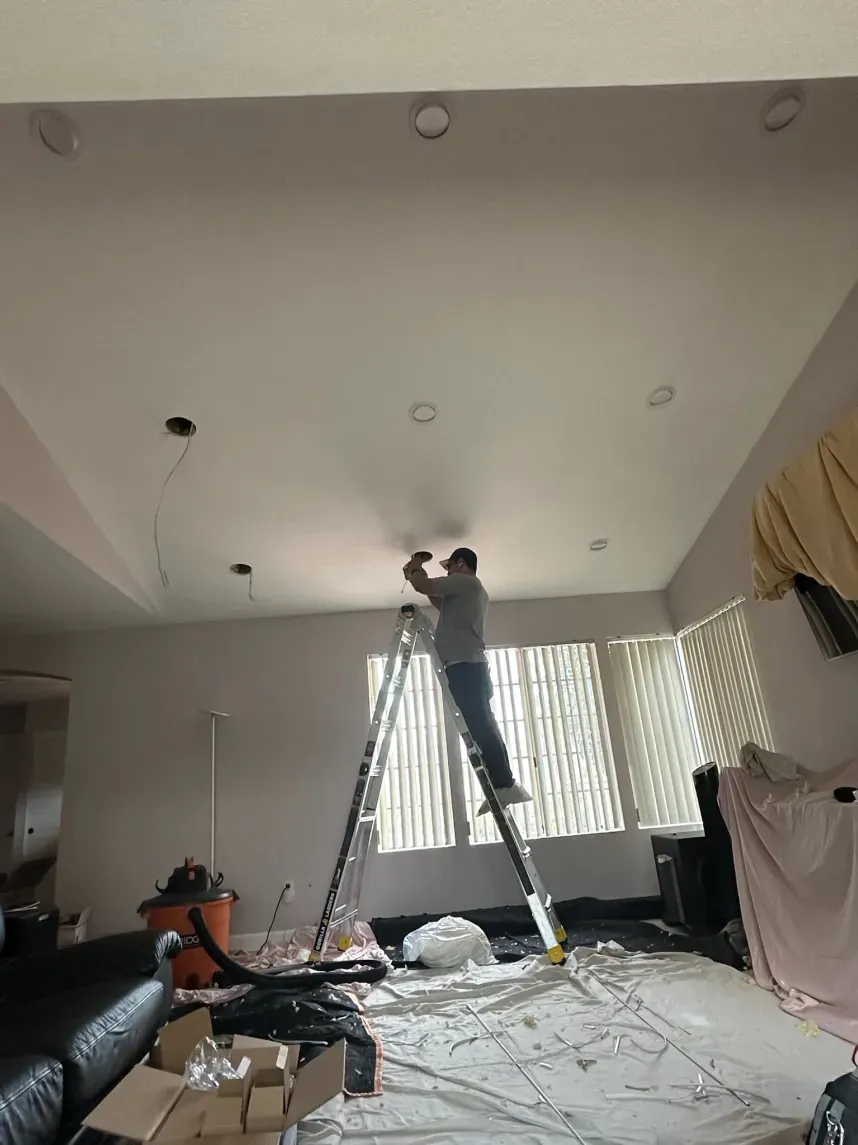 Person on ladder installing recessed lighting in a room with white walls and ceiling.