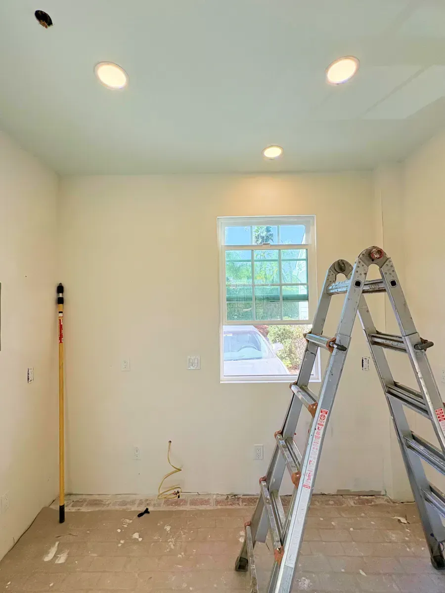 Room interior under construction with recessed lights, window, ladder, and paint supplies.