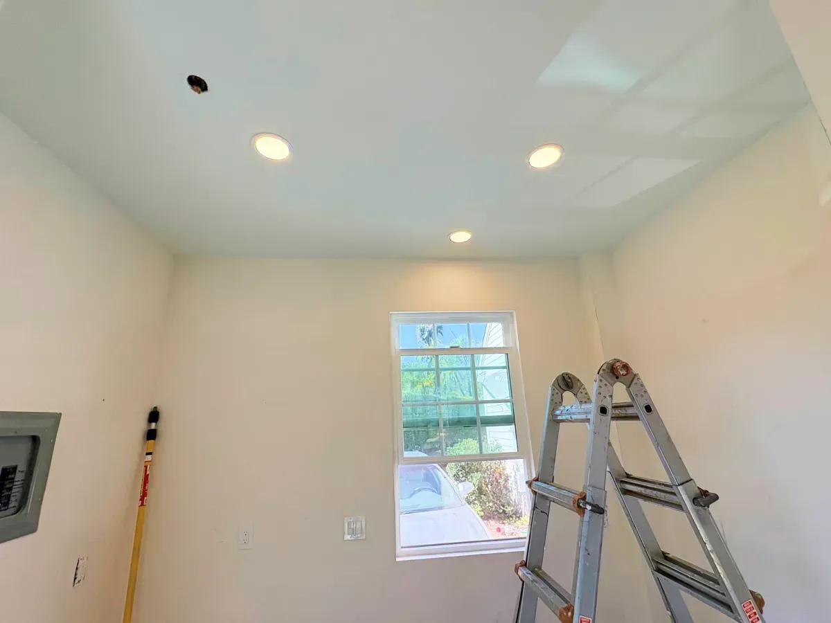 Interior view of a room under construction with recessed lighting and a window. A ladder stands by the window.