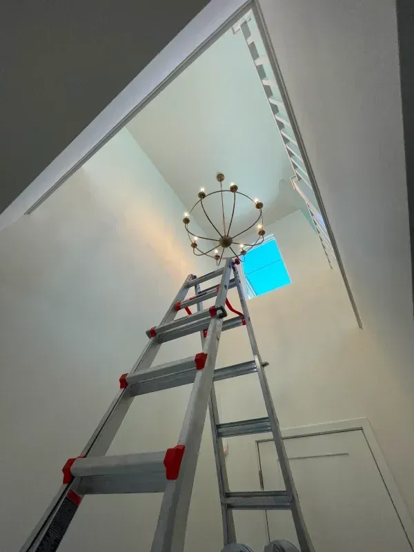 Ladder propped beneath a chandelier, reaching toward a ceiling. A light is on.