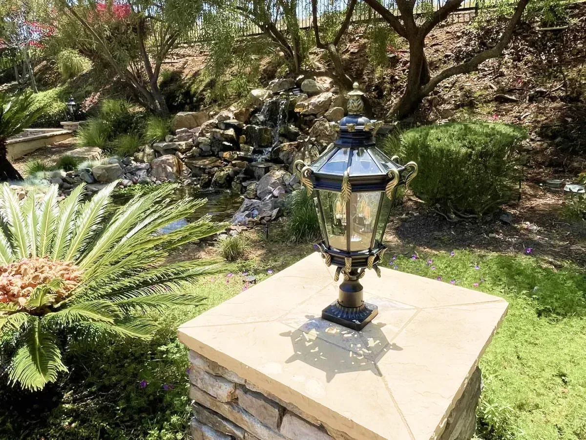 Decorative lamp on a stone pillar, set in a lush garden with a small waterfall in the background.
