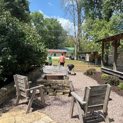 Two people grilling near a stone fire pit, two wooden chairs sit nearby. Sunny outdoor setting.